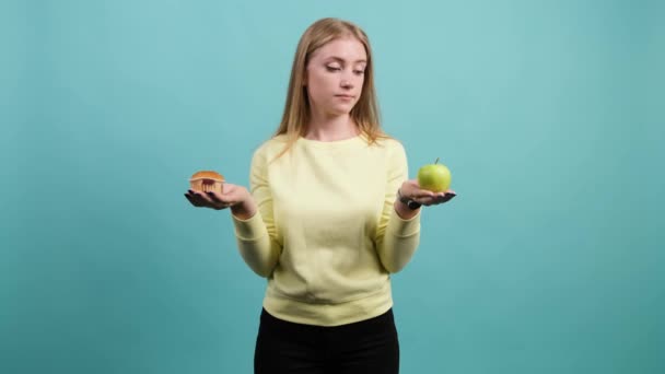 Młoda kobieta wybierając między jabłkiem a babeczką, dziewczyna jest zdezorientowana w swoim wyborze - Materiał filmowy, wideo