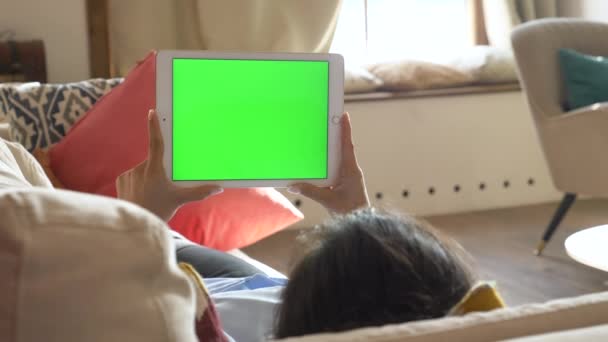 Mujer india joven sosteniendo una pantalla verde Chromakey Tablet en su mano, acostado en el sofá, trabajando en línea en una habitación brillante en casa - Imágenes, Vídeo