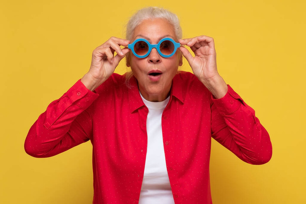Cheerful old woman wearing trendy sunglasses, isolated on yellow background laughing. - Фото, зображення