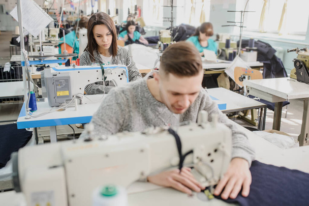 close up photo of a young man and other seamstresses sewing with sewing machine in a factory - Фото, изображение
