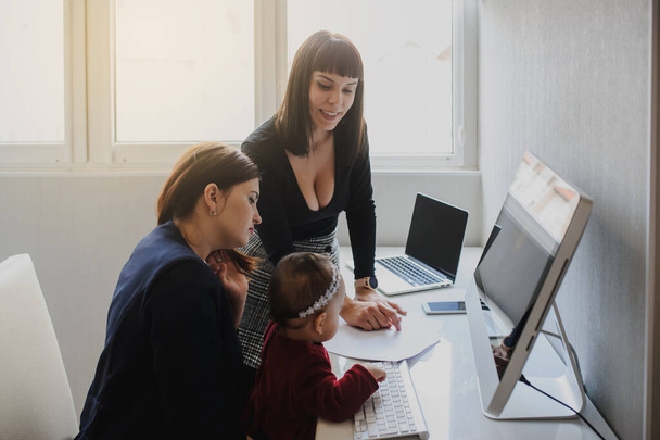 zbliżyć zdjęcie dwóch kobiet w stroju biurowym pracujących na komputerach i omawiających raport na podłodze; jedna z nich jest z dziewczynką w ramionach - Zdjęcie, obraz