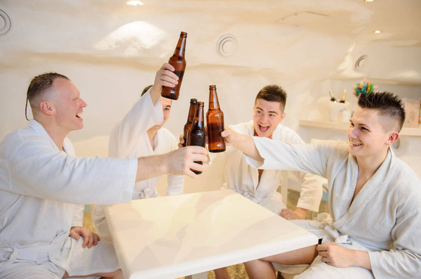 close up photo of 4 men in white gowns sitting around a table and drinking beer after sauna procedures - Fotó, kép