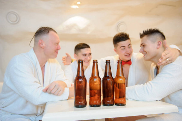 primer plano foto de 4 hombres en batas blancas sentados alrededor de una mesa y beber cerveza después de los procedimientos de sauna - Foto, imagen