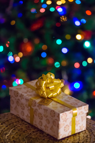 Belo presente de Natal com um arco de ouro sob a árvore contra um fundo de luzes bokeh multicoloridas desfocadas. Cartão postal, papel de parede, vertical - Foto, Imagem