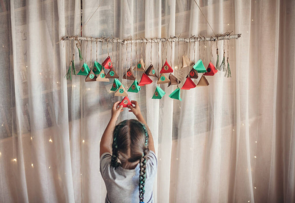 色の紙の三角形で手作りのアドベンチャーカレンダーを開くかわいい子供。お菓子や飴は枝に吊るされたカラフルな三角形に隠されています。子供のための季節の活動、家族の冬の休日 - 写真・画像
