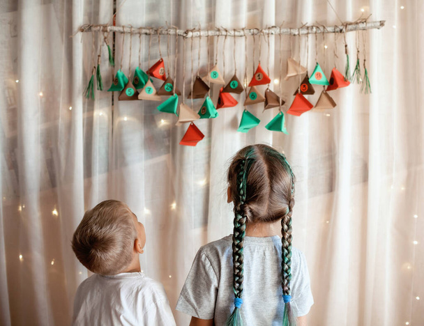 色の紙の三角形で手作りのアドベンチャーカレンダーを開くかわいい子供。お菓子や飴は枝に吊るされたカラフルな三角形に隠されています。子供のための季節の活動、家族の冬の休日 - 写真・画像