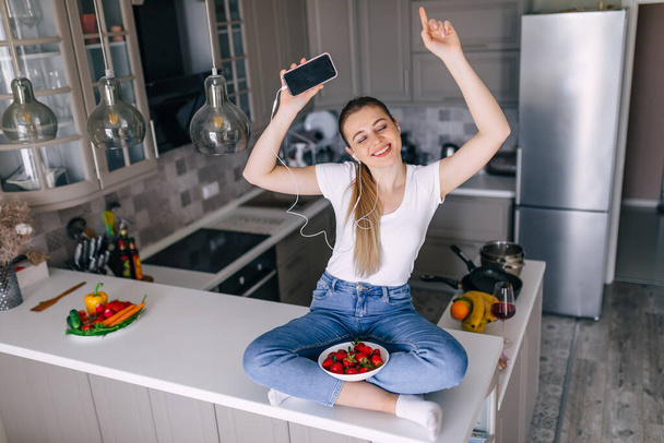 Image of young beautiful woman listening to music on mobile phone holding a plate of strawberries - Foto, imagen