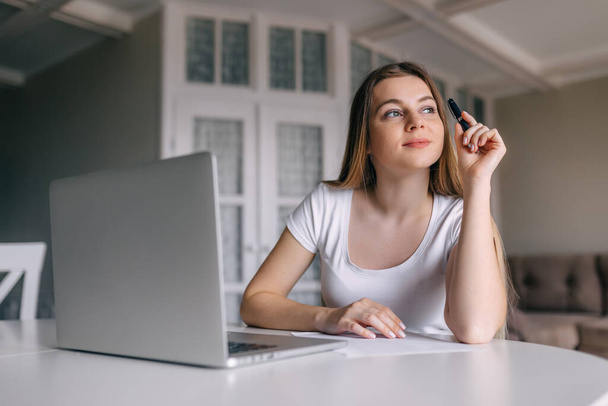 Single calm young woman in t-shirt and long hair holding pencil in hand while seated at desk in front of laptop computer in bright room - Foto, Bild