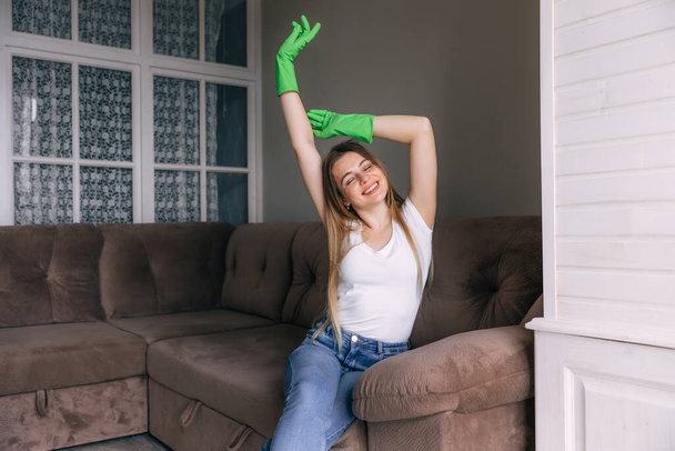 ευτυχισμένη νοικοκυρά κάθεται στον καναπέ φορώντας πράσινα γάντια μετά τον καθαρισμό του σπιτιού. - Φωτογραφία, εικόνα