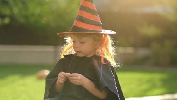 I bambini mangiano caramelle per Halloween. Ritratto di bambino caucasico in costume da pipistrello e cappello seduto a mangiare lecca-lecca dal secchio di zucca alla festa di Halloween - Filmati, video