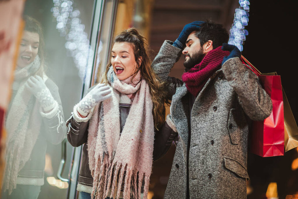 Pariskunta lahjakassin kanssa jouluvalojen taustalla kävellessä kaupungissa illalla. Ikkuna-ostokset. - Valokuva, kuva