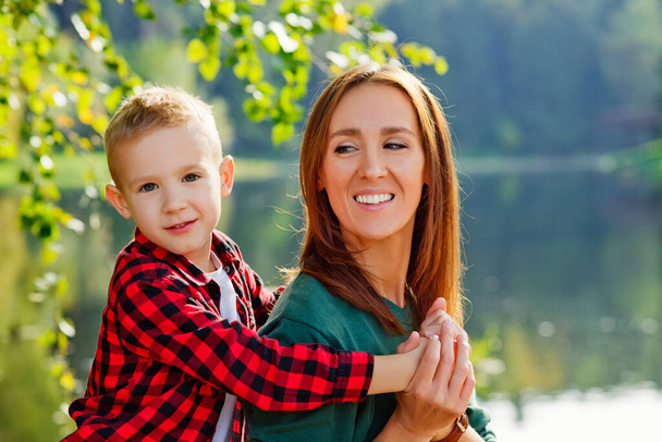 湖の背景に幸せな息子は首の後ろに若い笑顔の母親を抱擁。親、家族の日5月15日愛、親、子供の概念 - 写真・画像