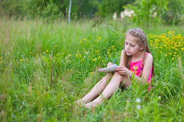 A young sad girl is sitting on a green grass in summer and writing something in a paper notebook - Foto, afbeelding
