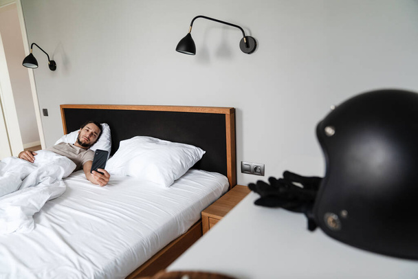 Усміхнений молодий чоловік використовує мобільний телефон, лежачи в ліжку вранці
 - Фото, зображення