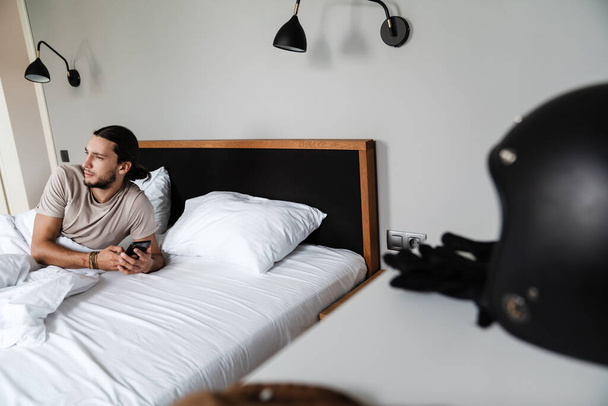 Smiling young man using mobile phone while laying in bed in the morning - Fotoğraf, Görsel