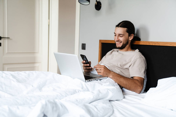 Jovem sorridente usando telefone celular e laptop enquanto deitado na cama pela manhã - Foto, Imagem