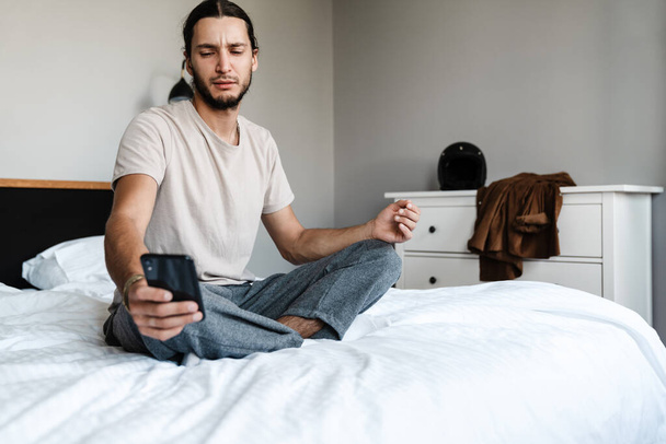 Upset young man looking at mobile phone while sitting on bed at home - Foto, Bild