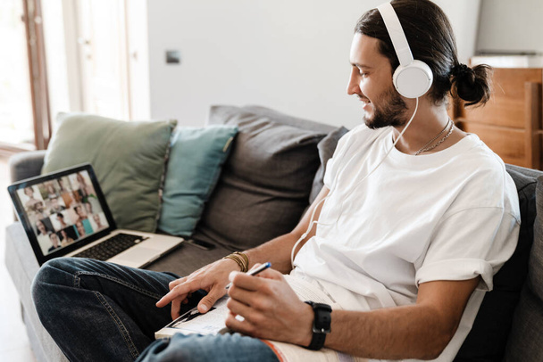 Confident young man wearing headphones having a video meeting via laptop while sitting on a couch at home - 写真・画像
