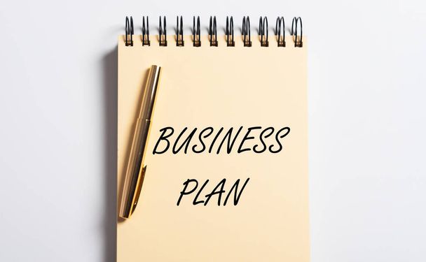 Businessplan-Schriftzug auf gelbem Spiralheft und goldenem Stift flach auf weißem Hochglanztisch, Draufsicht, Nahaufnahme - Foto, Bild