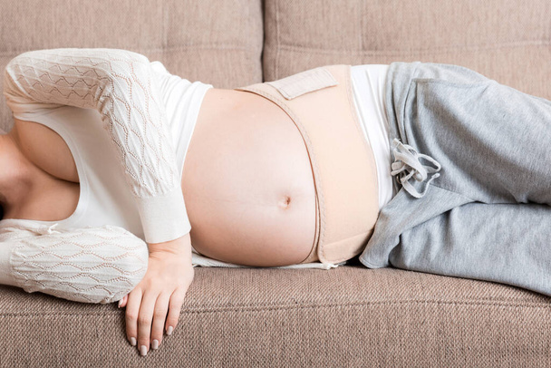 Terhes nő fekszik a kanapén a hasi kötésben. Anyasági egészségügyi ellátás koncepcióját támogató termékek. - Fotó, kép