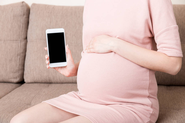 Schwangerschaft mit modernen Technologien. Schwangere Frau zeigt Handy mit leerem Displayauf dem heimischen Sofa. - Foto, Bild