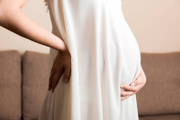 妊娠中の女性は自宅のソファで腰痛を抱えています。マタニティヘルスケアのコンセプトサポート製品. - 写真・画像
