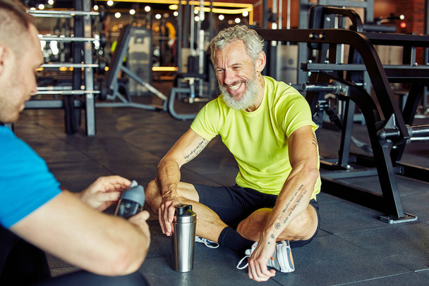 Ejercicio con entrenador personal. Feliz hombre de mediana edad discutiendo algo con instructor de fitness y sonriendo mientras están sentados juntos en el suelo en el gimnasio - Foto, imagen