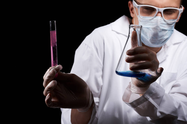 laboratory assistant in a medical mask and in a white coat with a liquid in a flask and a chemical on a black background - Photo, image
