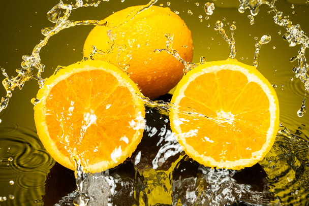 two oranges, whole and cut in half with drops and splashes of water on dark glass with gradient background and reflection - Фото, зображення