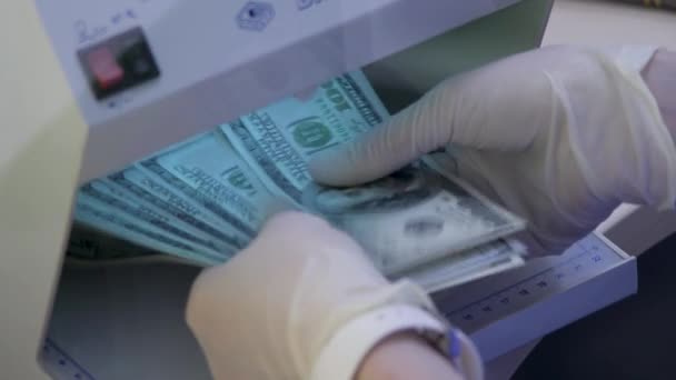 銀行の従業員または通貨交換事務員は、通貨検出器の紫外線下で100ドル札の現金を確認します。保護手袋の手紫外線検出器でドルのお金をチェック - 映像、動画