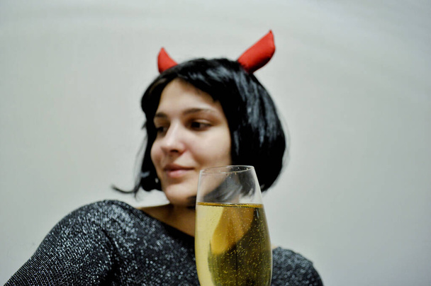 Mulher com cabelo preto curto com chifres vermelhos e uma taça de champanhe na mão sobre um fundo claro - Foto, Imagem