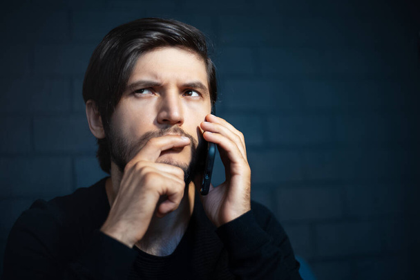 Close-up portrait of young thoughtful man, talking on smartphone. Background of black brick wall. - Photo, Image