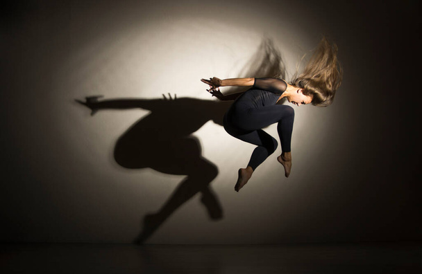 Girl in dark performs gymnastic jumping, on a white background there is a shadow from a shape. Studio photography. - Фото, изображение