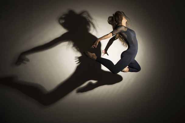 Girl in dark performs gymnastic jumping, on a white background there is a shadow from a shape. Studio photography. - Фото, изображение