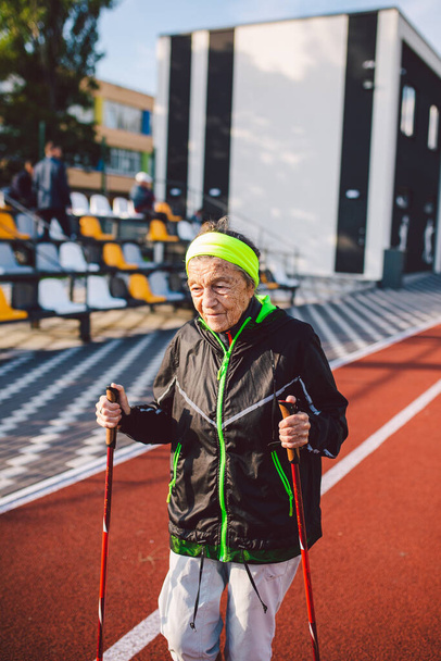 Yaşlı temasının geri kalanı aktif. Emeklilikte spor ve sağlık. Kafkas kırışıklıkları olan yaşlı bir kadın şehir stadyumunun dışında sopayla İskandinav yürüyüşü yapıyor.. - Fotoğraf, Görsel