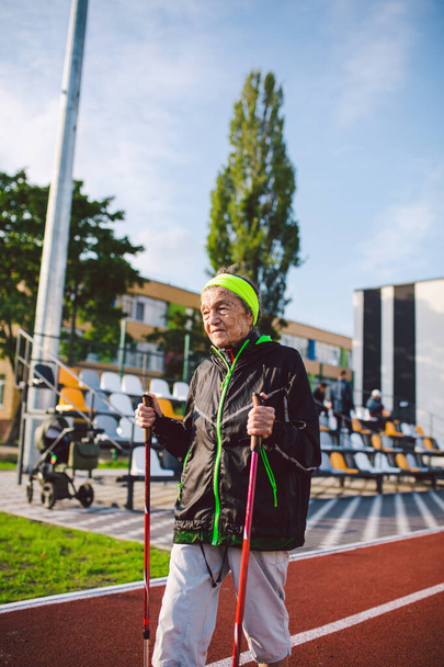 Aktivní starší ženy bělošky ve věku 90 let cvičí nordic walking s tyčemi na koleji s červenou gumovou vrstvou. Aktivní dovolenou. Hodná žena. Zdravý životní styl pro seniory. - Fotografie, Obrázek