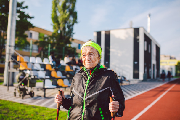 Активные пожилые кавказские пожилые женщины 90 лет практикуют нордическую ходьбу с лыжными палками на трассе с красным резиновым покрытием. Активный отдых. Подходящая женщина. Здоровый образ жизни для пожилых людей. - Фото, изображение