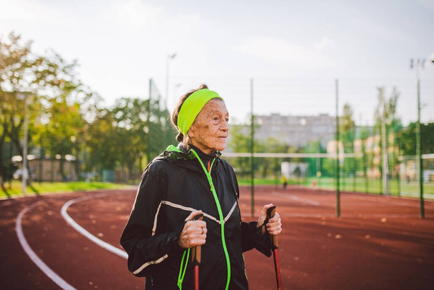 Starsza kobieta chodząca z kijkami na stadionie na czerwonej gumowej okładce. Starsza kobieta 88 lat robi ćwiczenia nordic walking na stadionie miejskim na torze biegowym. Koncepcja zdrowego stylu życia. - Zdjęcie, obraz