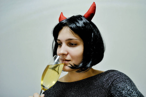 Woman with short black hair with red horns and a glass of champagne in her hand on a light background - Valokuva, kuva