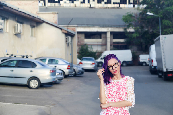 Joven mujer caucásica con el pelo morado y gafas, mirando a la cámara. Barrio industrial borroso fondo - Foto, imagen