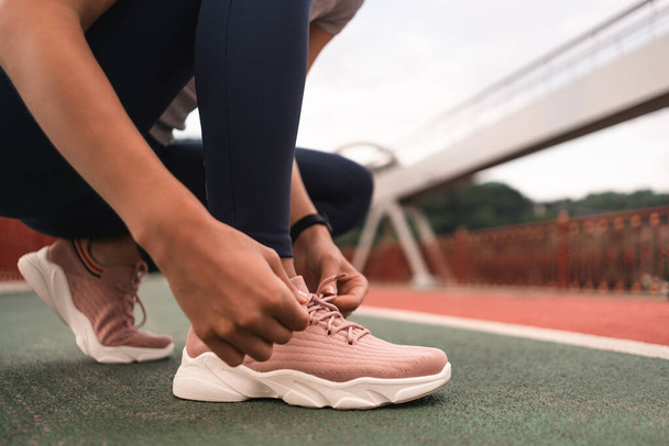 Cropped photo of hands of young woman lacing her pink sneakers while getting ready to jog on the bridge in the city. Focus on running shoes - Fotografie, Obrázek