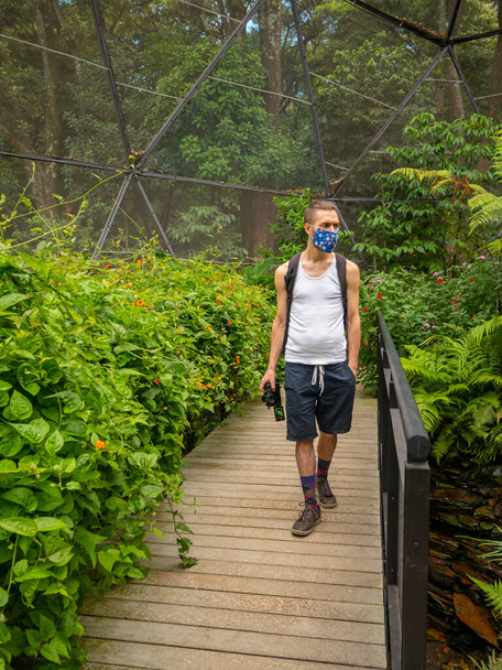 Hombre rubio caucásico con una máscara facial artesanal camina en el puente de madera en medio de la naturaleza - Foto, Imagen
