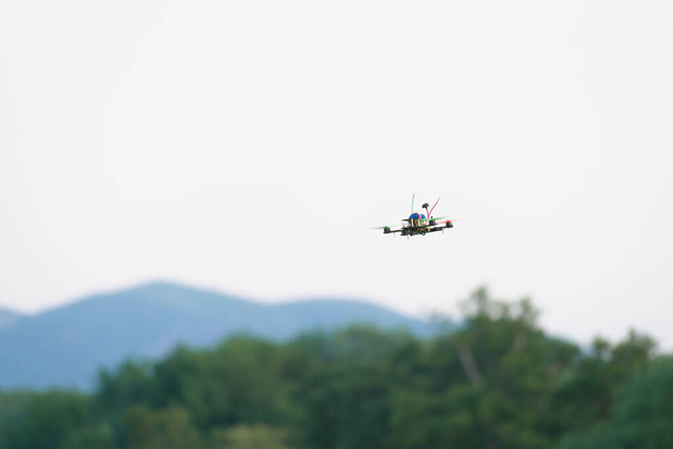 Πολύχρωμες προπέλες άρει αυτό το αγωνιστικό FPV quad copter drone μέσω του αέρα. Απλό λευκό φόντο του ουρανού με θολή λόφους και δέντρα κάτω - Φωτογραφία, εικόνα