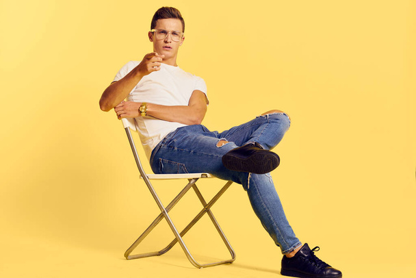 Homme mignon assis sur une chaise blanc t-shirt jeans style de vie moderne fond jaune - Photo, image