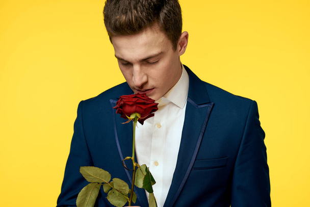 Mladý muž v klasickém obleku s červenou růží v ruce na žlutém pozadí emoce oříznuté pohled modelu - Fotografie, Obrázek