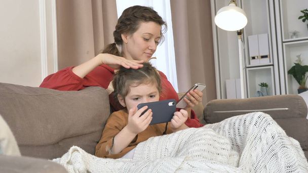 Mère caresse fille tenant des téléphones cellulaires sur le canapé dans la chambre - Photo, image