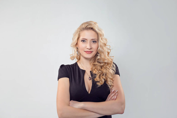 Happy and successful. Portrait of a happy beautiful smiling girl woman standing with arms folded isolated light grey white background. Caucasian Business person in black formal dress, long blond curly - Фото, изображение