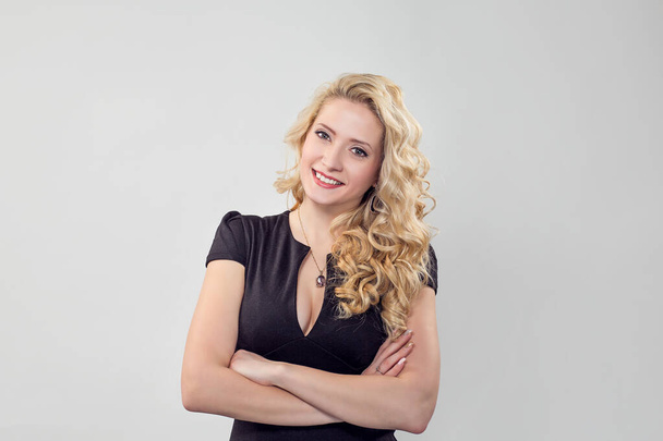 Front view of young smiling blonde in black dress standing looking at camera happy toothy smile. Caucasian Business person in black formal dress, long blond curly hair isolated on light grey gray wall - Foto, afbeelding