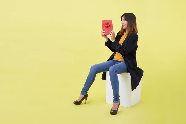 Счастливая привлекательная молодая женщина сидит на пустой пустой коробке с красной подарочной коробкой на желтом фоне - Фото, изображение