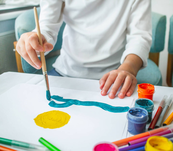 Το παιδί ζωγραφίζει τον ήλιο και τη θάλασσα σε γουάς πάνω σε ένα λευκό φύλλο χαρτί. Εκπαίδευση στο σπίτι. Εκπαίδευση σχεδίου. Παιδική ανεξαρτησία της τέχνης. - Φωτογραφία, εικόνα